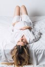 Сверху молодая нежная беременная женщина касается живота, лежа на кровати и счастливо улыбаясь — стоковое фото