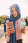 Araberinnen mit Kopftuch und Kopfhörern surfen auf dem Handy und hören Musik, während sie Kaffee zum Mitnehmen genießen, sonniger Tag in der Stadt — Stockfoto