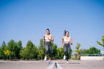 Повне тіло усміхнених спортивних близнюків у спортивному одязі, що бігають разом на асфальтній дорозі під час фітнес-тренувань проти зелених дерев — стокове фото