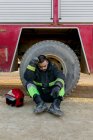 Виснажений чоловічий пожежник у формі сидить на землі біля пожежного двигуна біля червоного шолома, нахиляючи голову на руку — стокове фото