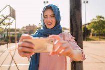 Очаровательная мусульманка в традиционном платке, стоящая на городской улице и стреляющая на смартфоне в солнечный день — стоковое фото
