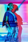 Vista lateral da moda jovem Dominicana millennial feminino com longas tranças afro em pé no chão e olhando para o lado enquanto ouve música em fones de ouvido no quarto com iluminação geométrica colorida — Fotografia de Stock