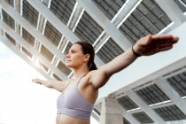 Mulher determinada em activewear com braços estendidos fazendo exercício de ioga na rua perto do painel fotovoltaico moderno durante o treinamento na cidade — Fotografia de Stock