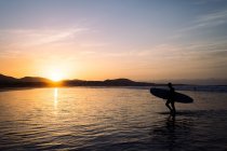 Seitenansicht der Silhouette eines anonymen Athleten mit Surfbrett, das im wogenden Meer gegen den Famara-Berg bei Sonnenuntergang auf Lanzarote spaziert — Stockfoto