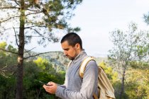 Vista lateral del joven viajero masculino étnico barbudo concentrado en ropa casual y mensajería de mochila en un teléfono inteligente de pie en un exuberante bosque verde durante el trekking en el valle montañoso - foto de stock