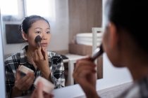 Vista lateral da jovem fêmea asiática em roupas casuais aplicando pó enquanto estava em frente ao espelho no quarto leve — Fotografia de Stock