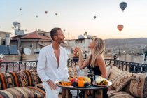 Пара держась за руки и глядя друг на друга, наслаждаясь романтическим ужином и кормя друг друга фруктами на террасе с воздушными шарами на заднем плане в Каппадокии — стоковое фото