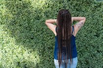 Вид сзади на анонимную латиноамериканку с татуировкой и длинными черными плетеными волосами во время стояния и размещения волос с руками рядом с зелеными растениями — стоковое фото