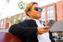 Cheerful young male in sunglasses and stylish outfit text messaging on cellphone while sitting on street near building in city — Stock Photo