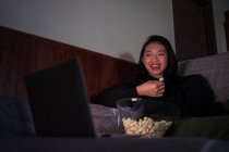 Young cheerful Asian female wearing casual clothes sitting on couch and eating popcorn at home watching movie on laptop — Stock Photo