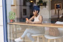 A través de la ventana de la joven mujer étnica con el pelo negro Afro en traje de moda y verdaderos auriculares inalámbricos felizmente mientras que tener una conversación de vídeo en el teléfono inteligente en la cafetería moderna - foto de stock