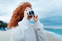 Positive ginger haired female in knitted sweater taking picture on retro photo camera on coast of sea — Stock Photo