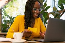 Positiva freelancer afroamericana con tarjeta de crédito sentada en la mesa con netbook mientras hace la compra en línea en la terraza en la cafetería - foto de stock