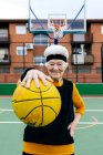 Положительная зрелая женщина в спортивной одежде и повязке смотрит в камеру, стоя с мячом в протянутой руке во время игры в баскетбол — стоковое фото