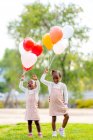 Longitud completa de hermanas pequeñas afroamericanas felices en vestidos similares de pie con globos de colores en las manos en la hierba verde en el parque a la luz del día - foto de stock