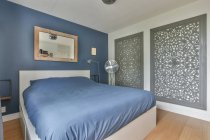 Удобная кровать с голубым одеялом размещена в стильной спальне с вентилятором и креативными декоративными элементами на стене в современной квартире — стоковое фото