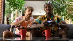 Diverse male friends with gamepads drinking beverages while sitting on couch and playing video game together in living room with green plants — Stock Photo