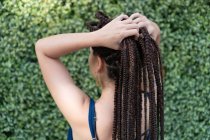 Back view of anonymous Hispanic female with tattoo and long black braided hair while standing and placing the hair with hands near green plants — Stock Photo