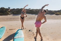 Corpo completo di coppia sportiva in costume da bagno guardarsi mentre si estende il corpo sulla spiaggia di sabbia soleggiata con tavole da surf — Foto stock