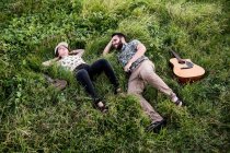 Happy friends musicians with guitar and ukulele sitting on green grass on coast near ocean in nature in daytime — Stock Photo