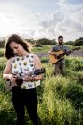 Mulher calma em roupas casuais tocando ukulele e de pé em campo verde contra amigo concentrado tocando guitarra na natureza em dia ensolarado — Fotografia de Stock