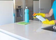 Crop anonymous woman in yellow rubber gloves spraying detergent on cloth while cleaning light kitchen — Stock Photo