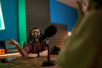 Jovem apresentador de rádio masculino em camisa quadriculada e fones de ouvido sentado à mesa com microfone e se comunicando com colega anônimo durante gravação de podcast em estúdio — Fotografia de Stock