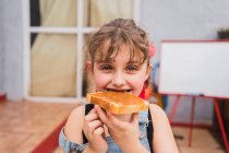 Nettes Mädchen in Jeans schaut in die Kamera, während es zu Hause frisches Brot mit süßer Marmelade isst — Stockfoto
