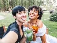 Encantada multirracial mejores amigas con vasos de cócteles escalofriantes en el parque de verano y tomar selfie en el teléfono inteligente mientras sobresale la lengua - foto de stock