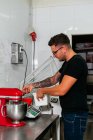 Side view of young tattooed male confectioner in casual clothes and eyeglasses weighing flour on electronic scale standing at table with mixer in kitchen — Stock Photo