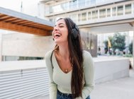 Весела жінка з довгим коричневим волоссям в повсякденному одязі та навушниках слухає музику та сміється на вулиці міста в денний час — стокове фото