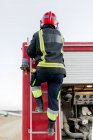 Вид сзади на анонимного пожарного в красном защитном шлеме и форме, стоящего на пожарной лестнице и смотрящего вдаль днем — стоковое фото