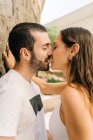 Vista lateral de la joven pareja étnica enamorada en ropa casual abrazándose y besándose de pie cerca de la pared de piedra en la calle de la ciudad - foto de stock