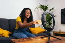 Mujer afroamericana en ropa de mezclilla sonriendo mientras está sentada en el sofá y tocando el teléfono inteligente en la lámpara de anillo brillante - foto de stock