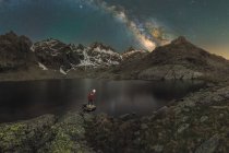 Back view of unrecognizable male tourist with torch admiring snowy mounts reflecting in water under starry sky at night — Stock Photo