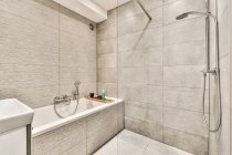 Contemporary shower room interior with bathtub and gray tiled wall against washbasin in light house — Stock Photo
