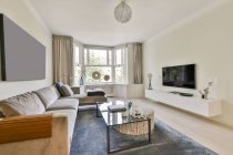 Comodo divano situato sotto pittura bianca vicino a tavolo di vetro e sgabello in luce elegante soggiorno — Foto stock