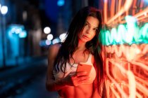 Текстовые сообщения для женщин на мобильном телефоне, стоя возле здания с светящимися неоновыми огнями на вечерней улице на размытом фоне — стоковое фото