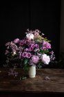 Букет свіжих барвистих півоній і хризантем у білій вазі, розміщеній на дерев'яному столі в темній кімнаті — стокове фото