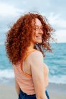 Vista posteriore di femmina deliziata con ricci lunghi capelli rossicci in piedi guardando oltre la spalla alla fotocamera contro il mare blu — Foto stock