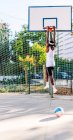 Back view of anonymous African American male basketball player hanging on metal hoop on playground in summer — Stock Photo