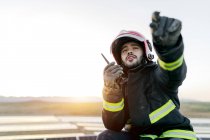 Позитивный молодой бородатый пожарный мужчина в шлеме и защитных перчатках и одежде, держа рацию в поднятой руке и мирно глядя в сторону на размытом фоне днем — стоковое фото