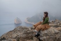Повний вид збоку тіла мрійливої жінки в старомодному одязі, що сидить на краю кам'яної скелі біля моря в туманній погоді — стокове фото