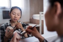 Vue latérale de la jeune femme asiatique en vêtements décontractés appliquant de la poudre tout en se tenant devant le miroir dans la chambre à coucher lumineuse — Photo de stock