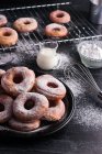 Солодкі смажені пончики подаються на тарілці біля металевої стійки для охолодження та глечика молока на чорному безладному столі з цукровою пудрою — стокове фото
