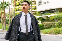 Хорошо одетый молодой азиатский предприниматель в галстуке смотрит в сторону, прогуливаясь по дороге против современных зданий в городе — стоковое фото