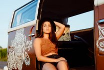 Menina morena atraente com chapéu sentado no chão de uma van vintage em um dia ensolarado — Fotografia de Stock