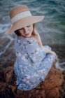 Charming young female in summer dress and hat sitting on rocky seashore with closed eyes in summer evening — Stock Photo