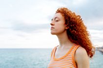 Спокойная женщина с вьющимися рыжими волосами наслаждается ветреной погодой на берегу бурлящего моря — стоковое фото