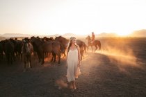 Donna bionda in abito bianco guardando lontano con mandria di cavalli in campo sotto il tramonto — Foto stock